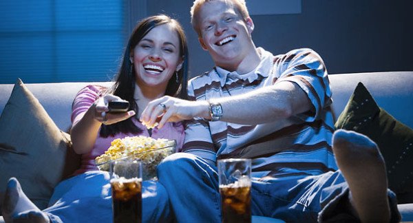 Couple sitting on sofa watching television with bowl of popcorn smiling